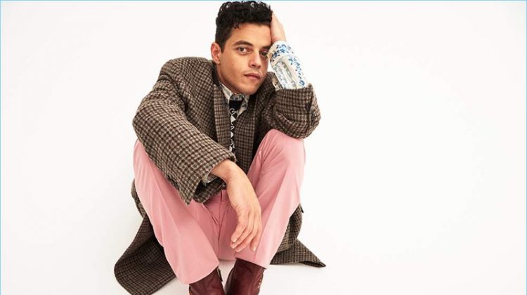 Rami Malek wears a Balenciaga checked coat with a shirt, boots, and sweater vest by Gucci. He also dons pink Alexander McQueen trousers.