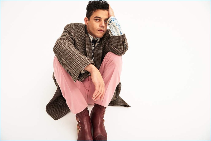 Rami Malek wears a Balenciaga checked coat with a shirt, boots, and sweater vest by Gucci. He also dons pink Alexander McQueen trousers.