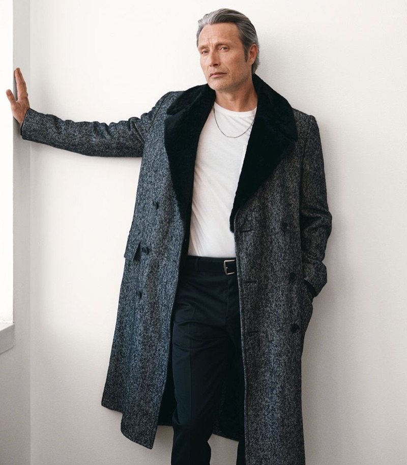 Connecting with Mr Porter, Mads Mikkelsen sports a Dunhill herringbone coat, Tom Ford t-shirt, Berluti trousers, Loro Piana suede belt, and Saint Laurent necklace.