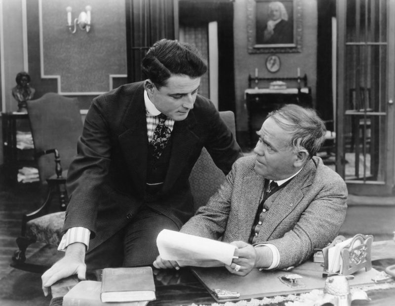 Two coworkers are going over paperwork at the moment. Men's hairstyles from the 1920s include a side part and a combover, which were both popular at the time.