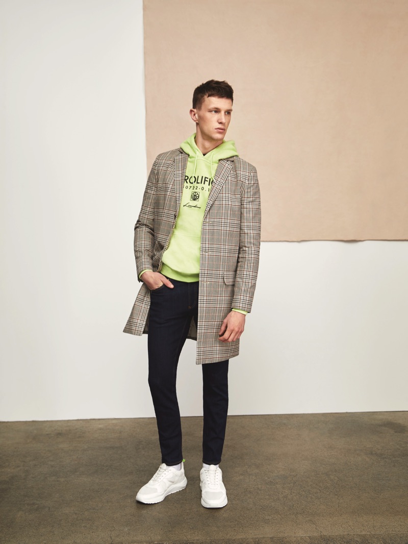 Donning a check River Island coat, Jack Buchanan also wears a bright lime green hoodie and slim-fit jeans with white sneakers.
