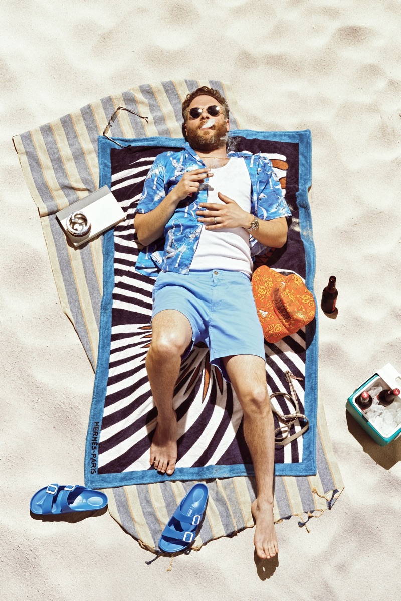 Relaxing on an Hermès beach towel, Seth Rogen wears a Dior Men shirt, Calvin Klein tank, POLO Ralph Lauren shorts, Birkenstock sandals, and Garrett Leight sunglasses.