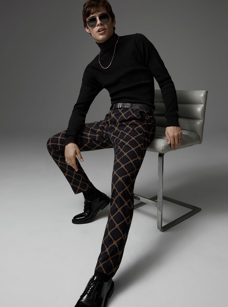 Model Louis Baines sports a LE 31 turtleneck sweater with ornamental pattern pants, a leather belt, and Ray-Ban sunglasses.