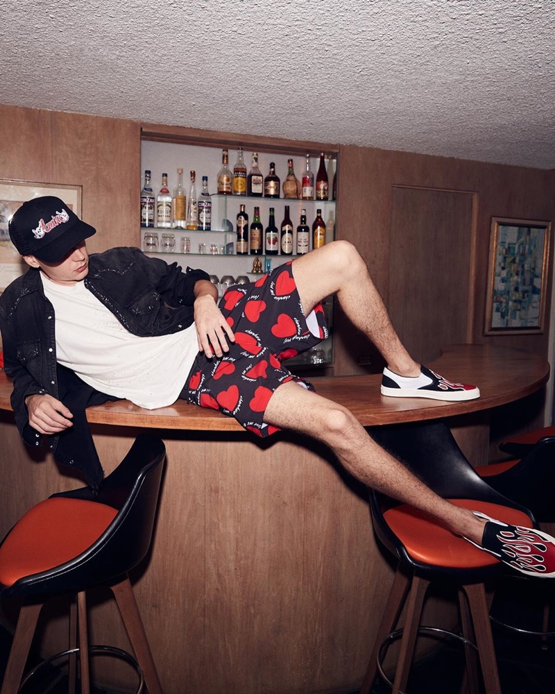 Relaxing on a bar top, Yuri Pleskun models an AMIRI Motley Crue trucker hat $275, heart print swim trunks $390, flame graphic slip-on shoes $595, studded denim shirt $950, and Shotgun tee $325.