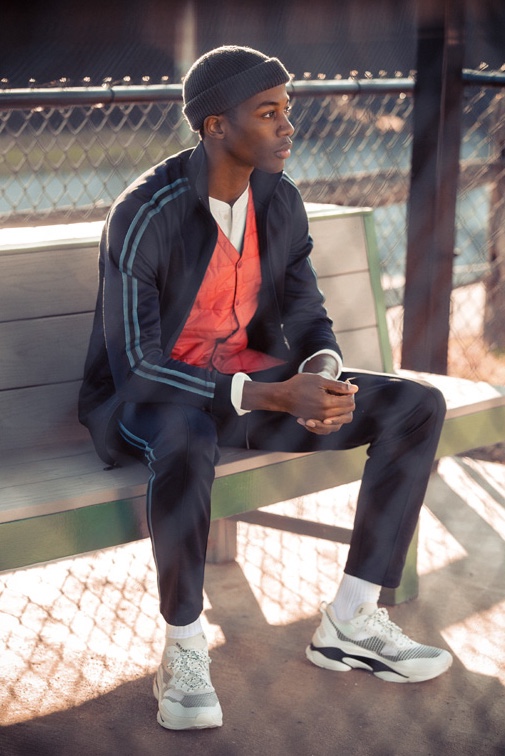 Magor Meng wears a Vince track jacket and pants with a shawl collar henley. He also sports a Gramicci Japan padded quilted vest, Brandblack Specter sneakers, and Filson watch cap.