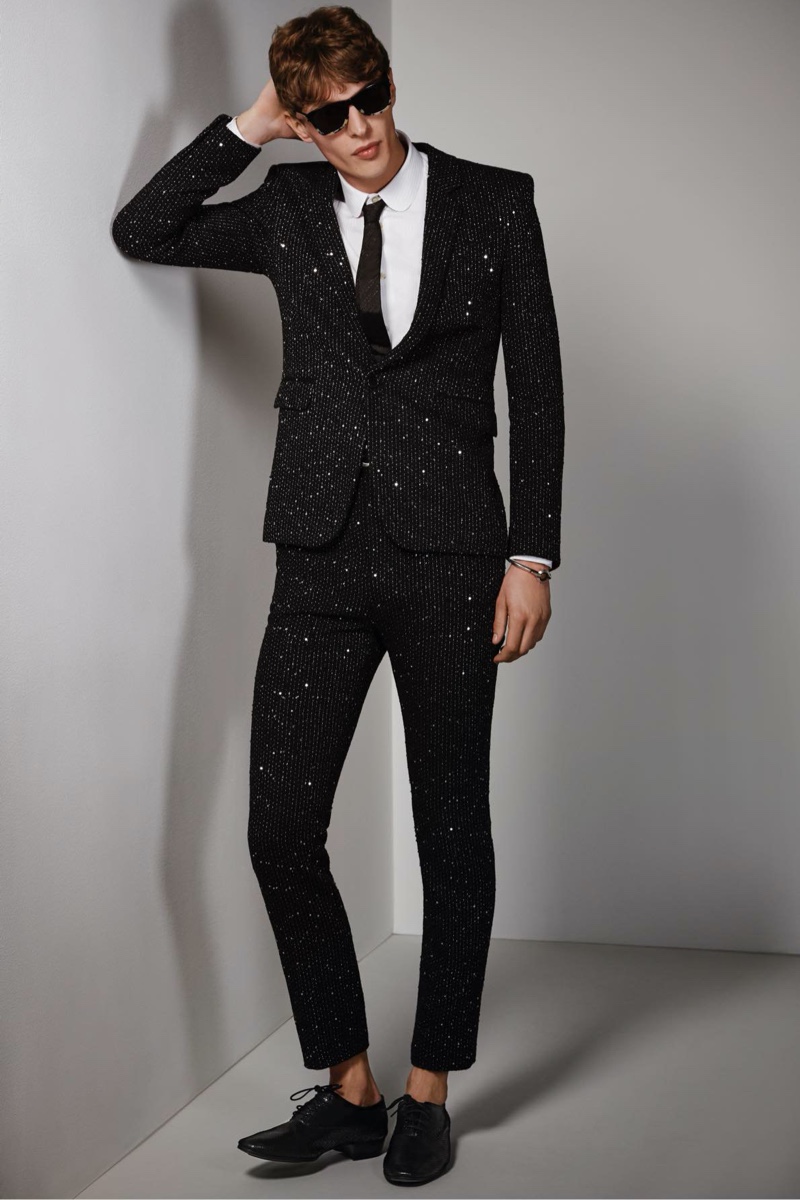 Tim Schuhmacher wears a twill bouclé sequin jacket and pants with a shirt, tie, sunglasses, bracelet, and patent leather oxfords by Saint Laurent.