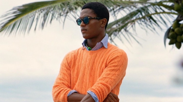 Hamid Onifade sports a bright orange cable-knit sweater with a striped oxford shirt and frayed white shorts by POLO Ralph Lauren.
