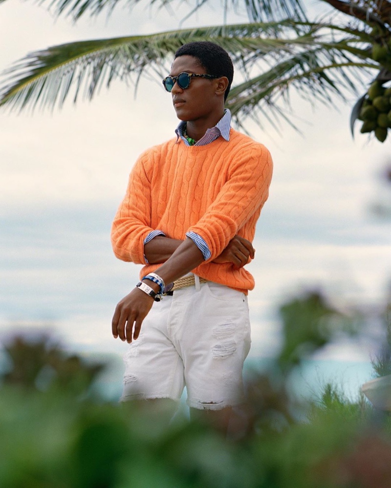 Hamid Onifade sports a bright orange cable-knit sweater with a striped oxford shirt and frayed white shorts by POLO Ralph Lauren.