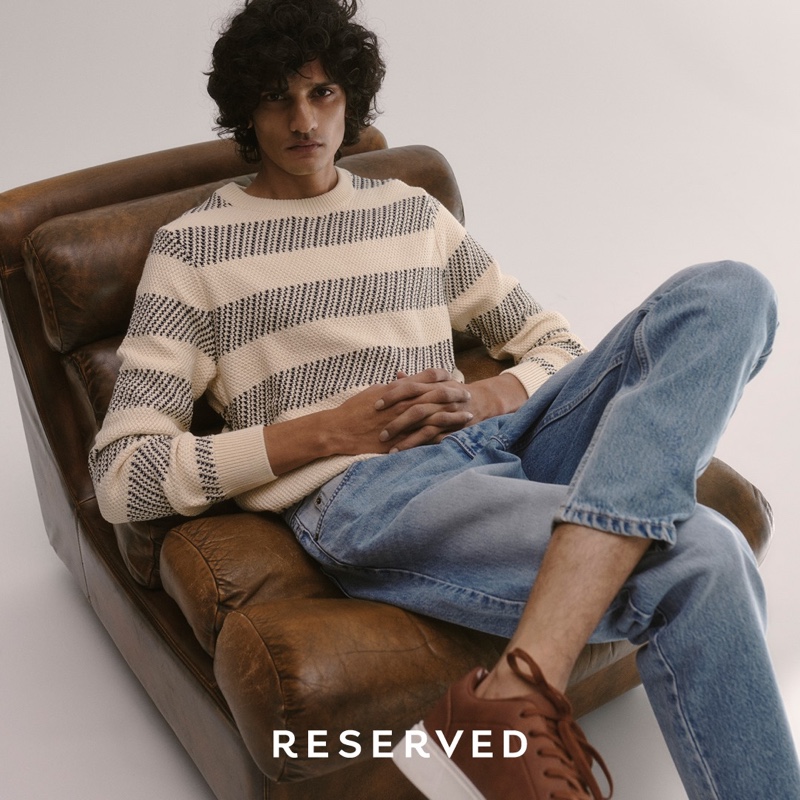 Reclining in a leather chair, Mustafa Dawood goes casual in a striped sweater and light wash denim jeans from Reserved.