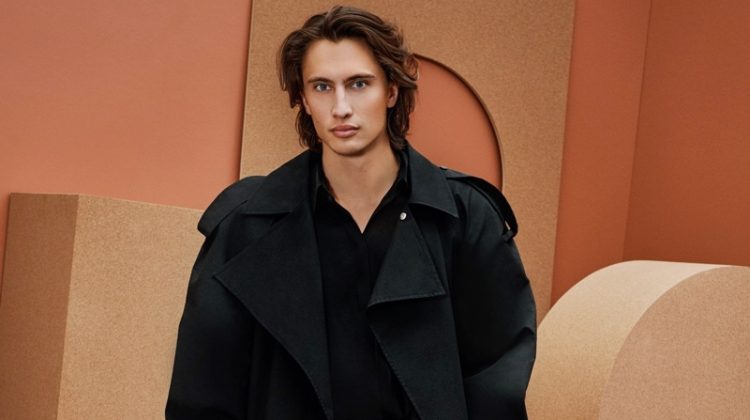 Front and center, James Turlington models a trench coat, long-sleeve shirt, leather shorts, and sandals from Bottega Veneta for Holt Renfrew.