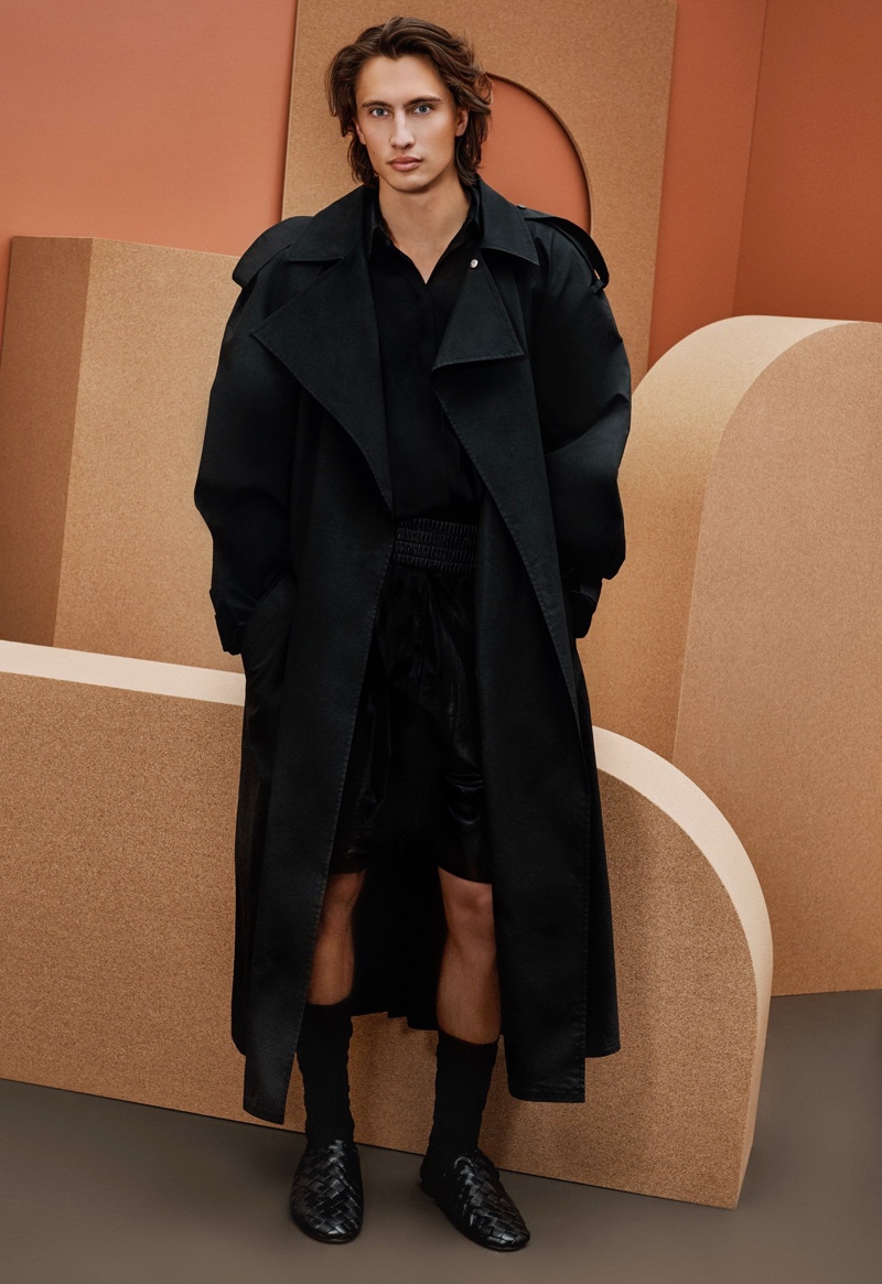 Front and center, James Turlington models a trench coat, long-sleeve shirt, leather shorts, and sandals from Bottega Veneta for Holt Renfrew.