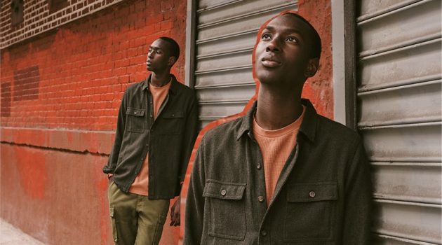 East Dane Proposes Casual Essentials with Fall Style Guide Fall Layers: Embracing autumnal tones, Macky Dabo layers in menswear from East Dane. Posing against a colorful brick wall and metal door, Macky wears a Banks Journal brushed herringbone shirt jacket and waffle fleece sweatshirt. He also models Alex Mill cargo pants with Birkenstock Soft Arizona suede sandals.