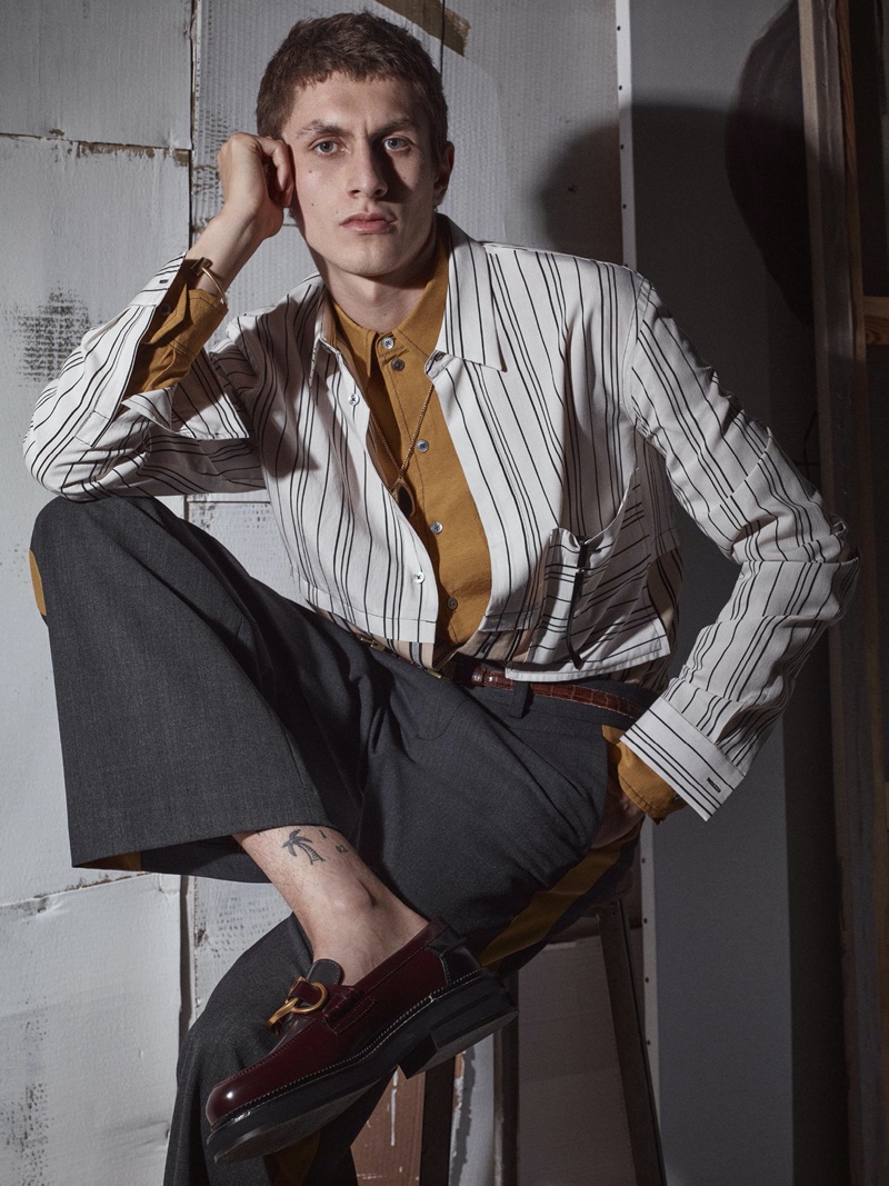 Henry Kitcher fronts Zara's fall-winter 2020 men's campaign. Sitting for a portrait, the top model wears a mixed stripe shirt over a button-down with trousers. A brown leather belt and loafers bring his look together.