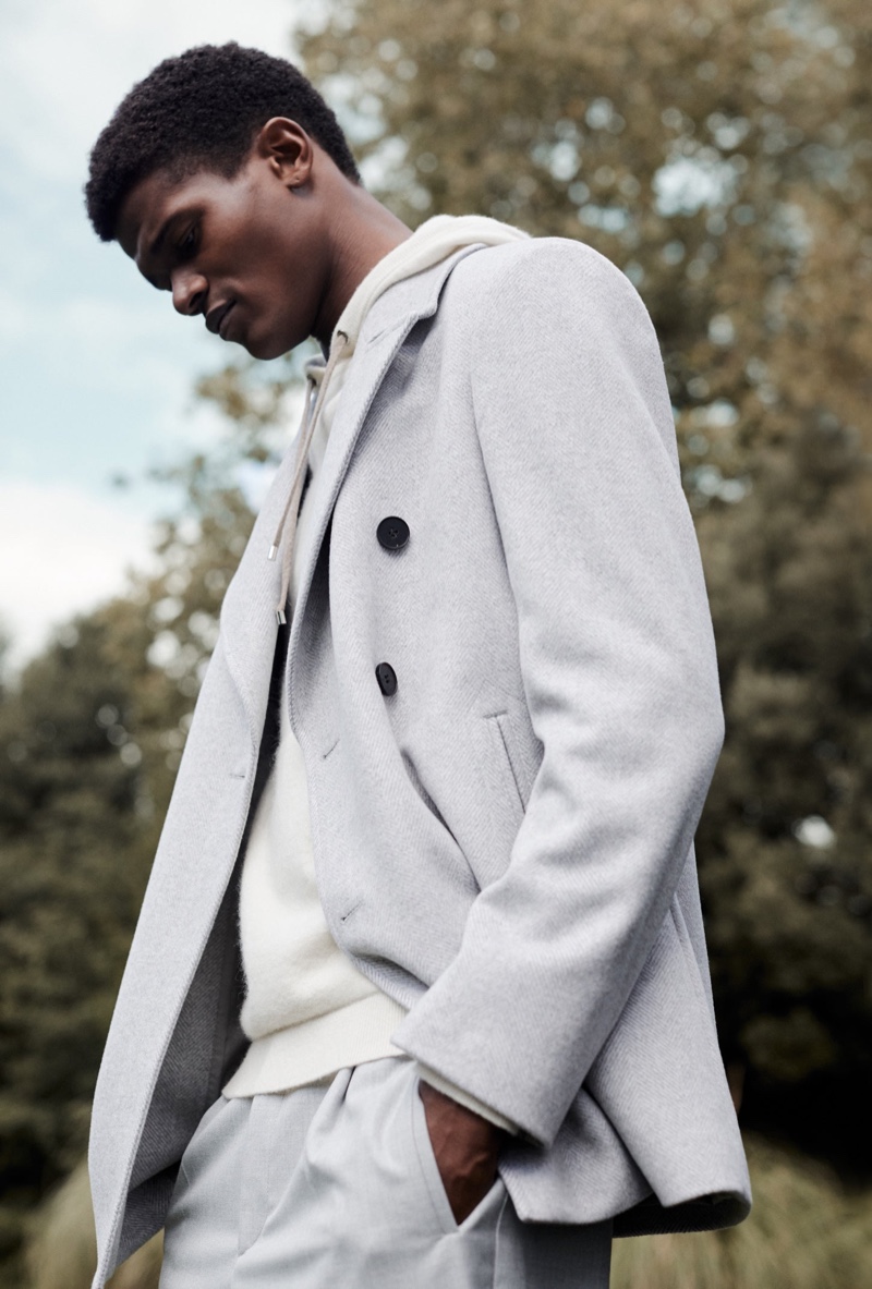 Delivering a side profile, O'Shea Robertson models a double-breasted coat with a hoodie and trousers for Reiss' fall-winter 2020 campaign.