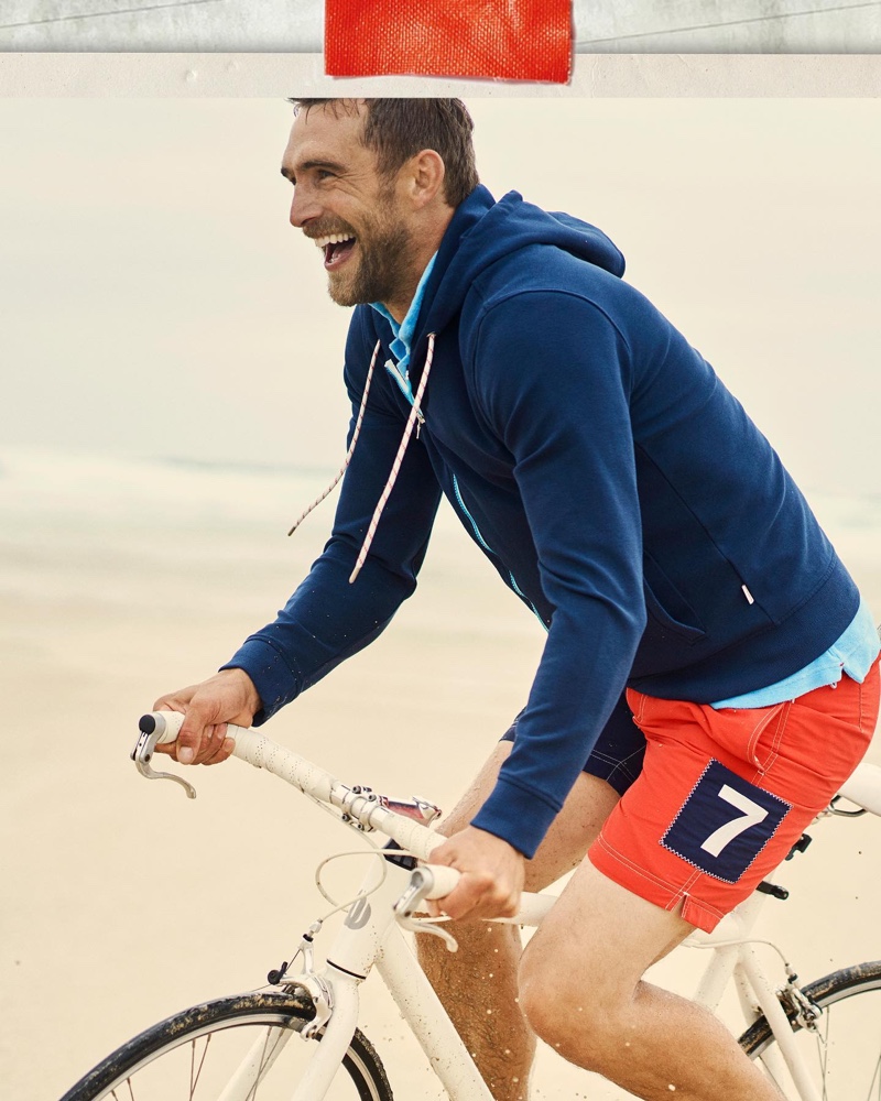 On the move, Will Chalker is all smiles in a navy zip-thru hooded sweatshirt and mid-length swim shorts from Orlebar Brown.