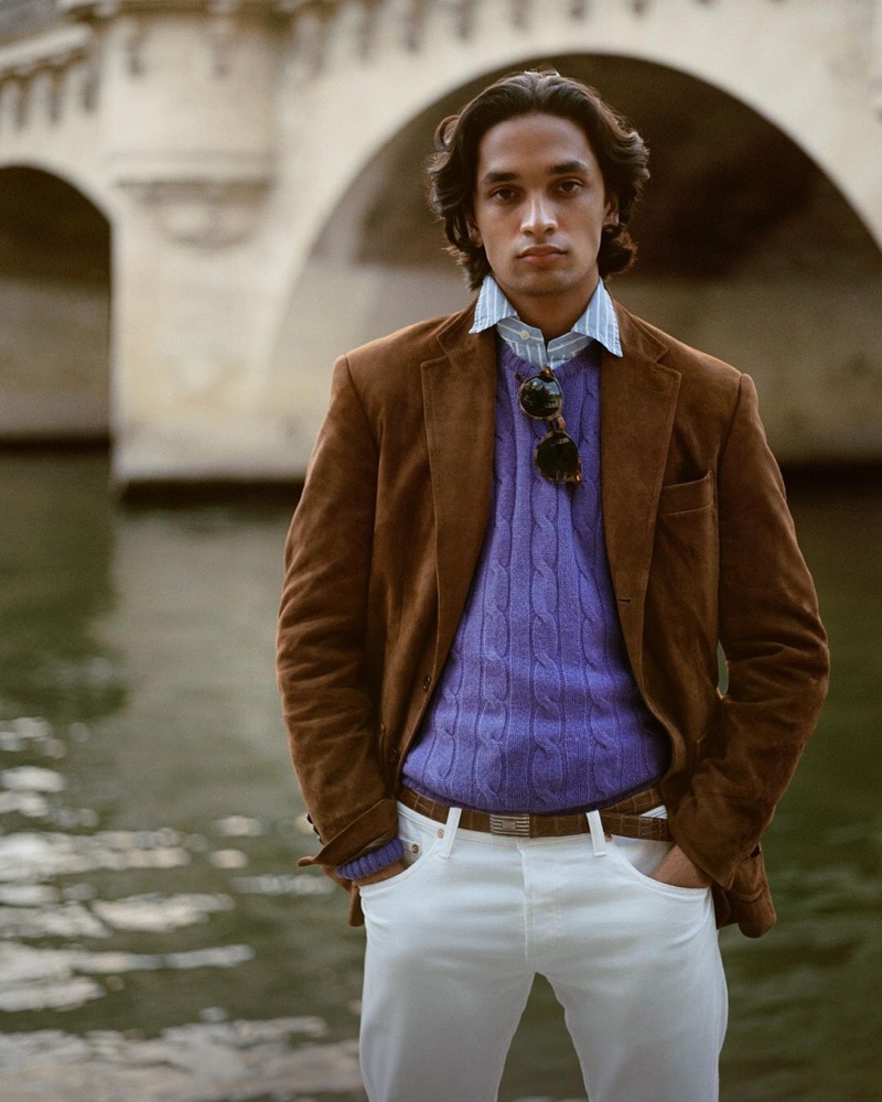 Model Sanjay Apavou dons a purple cable-knit sweater with a striped oxford shirt, brown suede jacket, and white pants from POLO Ralph Lauren.