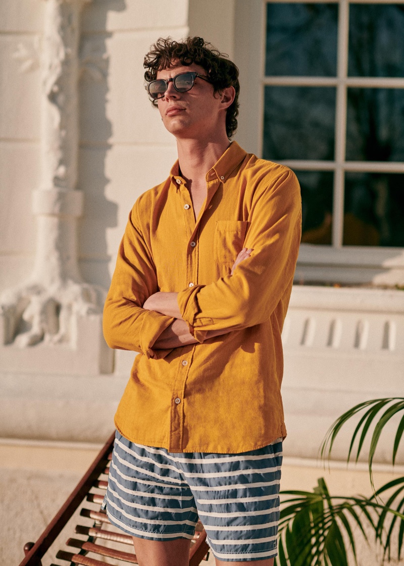 Dressing for warm weather, Andrea Quaranta models a mustard yellow shirt with striped swim shorts and stylish sunglasses by Octobre.