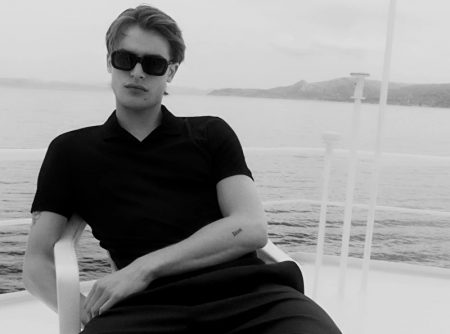 Yachting, Parker Van Noord relaxes in a black open collar polo shirt, drawstring wide-leg pants, and strap sandals by COS.