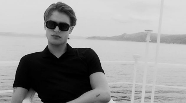 Parker Van Noord Dons High Summer Styles from COS Yachting, Parker Van Noord relaxes in a black open collar polo shirt, drawstring wide-leg pants, and strap sandals by COS.