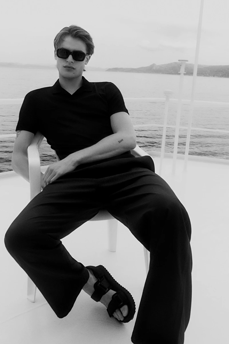 Yachting, Parker Van Noord relaxes in a black open collar polo shirt, drawstring wide-leg pants, and strap sandals by COS.