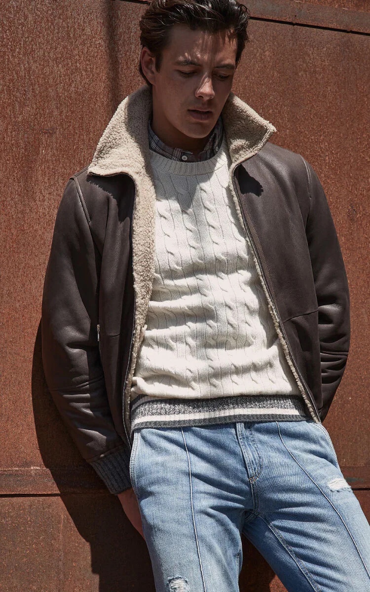 Harry Gozzett wears a leather jacket with a cable-knit sweater, plaid shirt, and jeans by Brunello Cucinelli.