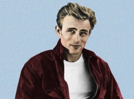 James Dean appears in a promotional image for Rebel Without a Cause (1955). The actor sports an iconic look consisting of a red Harrington jacket, white t-shirt, and Levi's jeans.