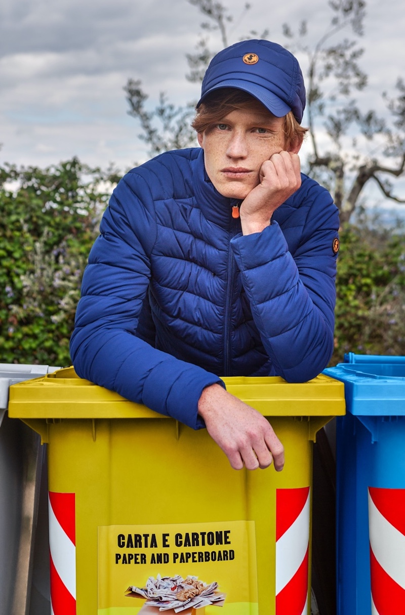 Sporting a matching blue cap and quilted jacket, model Christian Aneris fronts Save The Duck's new "Love Recycle" campaign for fall. Christian poses in a yellow recycling bin, reinforcing Save The Duck's advocacy for the environment.