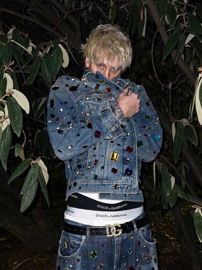 Hiding from the camera, Machine Gun Kelly rocks a denim jacket adorned with studs and colored crystals from Dolce & Gabbana's spring-summer 2022 collection. He also sports matching jeans, a DJ logo belt, and Dolce & Gabbana underwear.