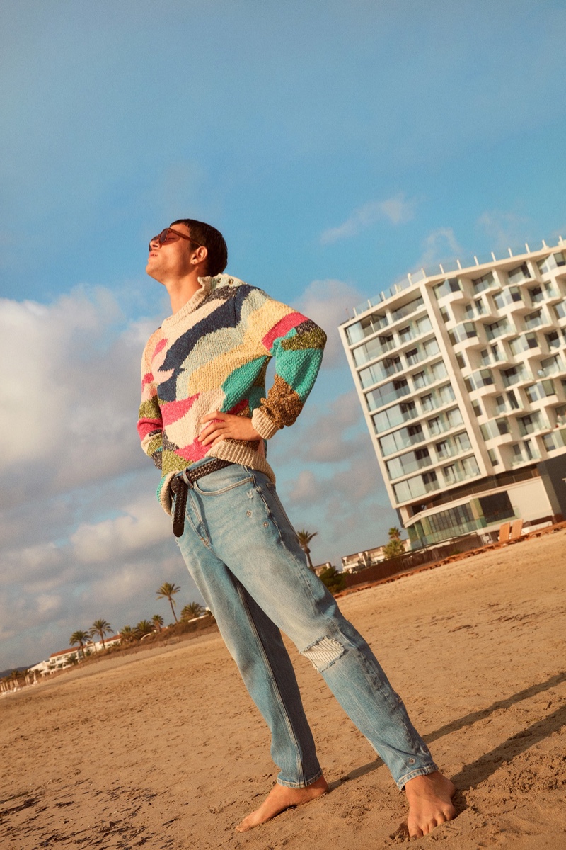 Hitting the beach, Luka Isaac wears a multi-colored sweater with denim jeans from Scotch & Soda's spring-summer 2022 collection for men.