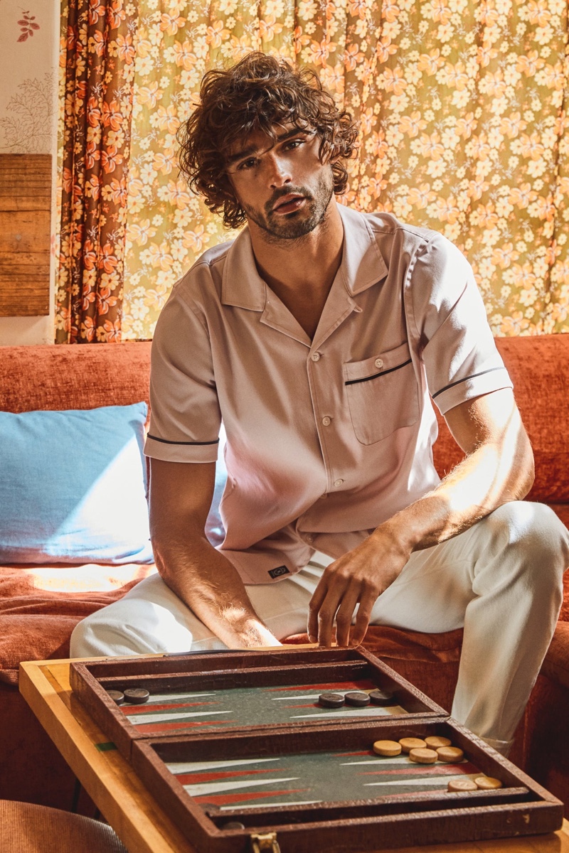 Playing a game of backgammon, Marlon Teixeira sports a Todd Snyder Japanese piped rayon shirt in pink with slim-fit 5-pocket chinos in manor grey.