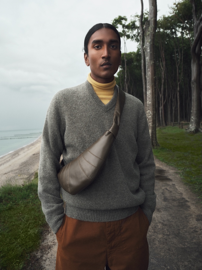 A smart vision, Francois Delacroix dons a v-neck sweater with straight-leg canvas pants, a yellow cotton jersey turtleneck top, and a croissant small leather shoulder bag from the LEMAIRE x Mytheresa collection.