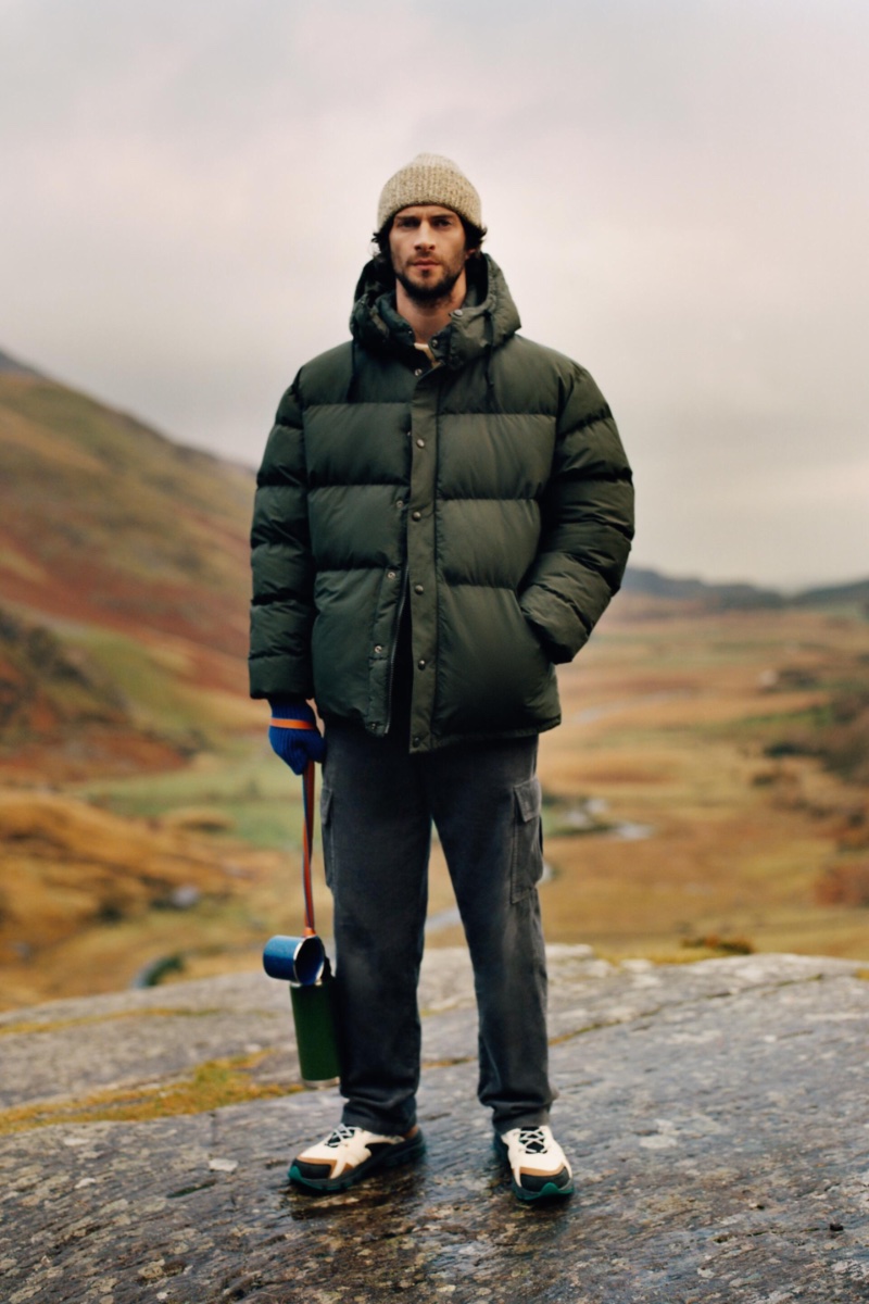 In front and center, Matthew Bell dons a hooded puffer jacket with cargo pants, a cotton knit beanie, and trekking high top sneakers. 