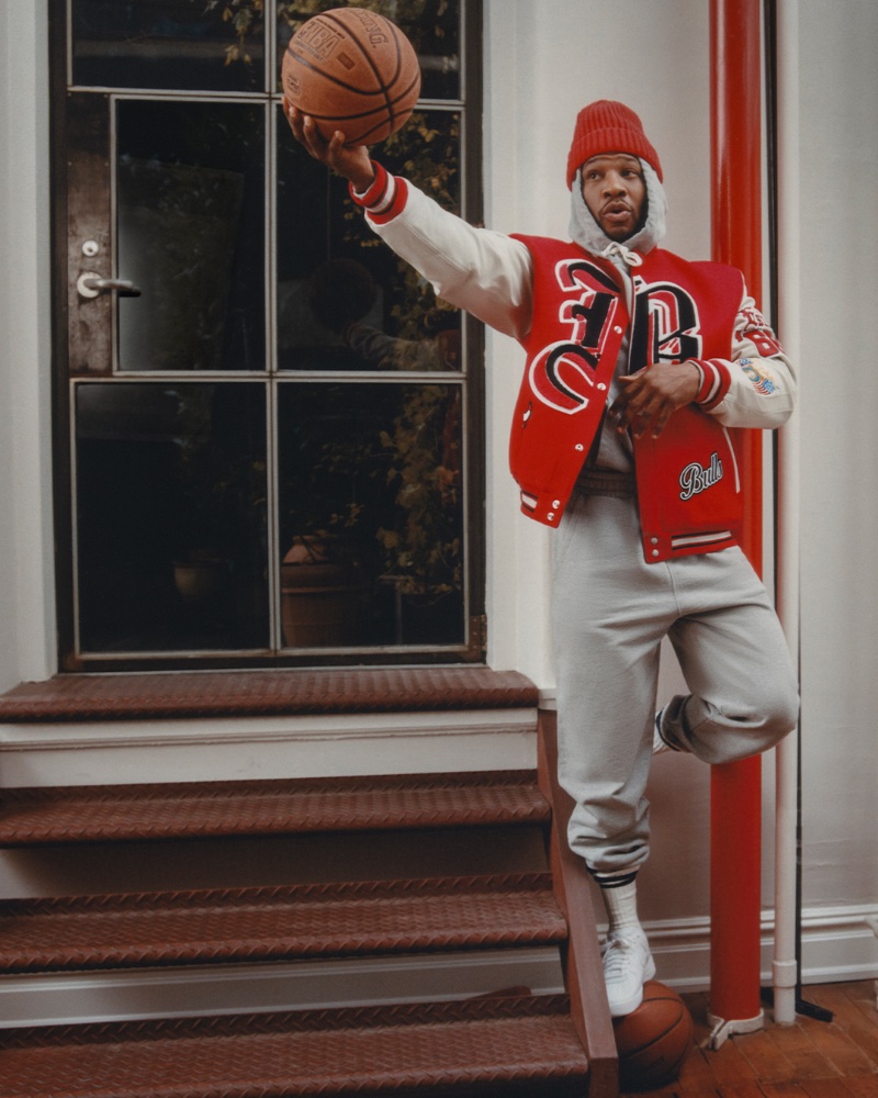 Styled for a sports day, Jonathan Majors wears a Jeff Hamilton Bulls varsity jacket, CDLP hoodie, and sweats. Beams Plus socks, a Drake's cashmere beanie, and Nike Air Force 1 '07 Fresh sneakers complete his outfit. 