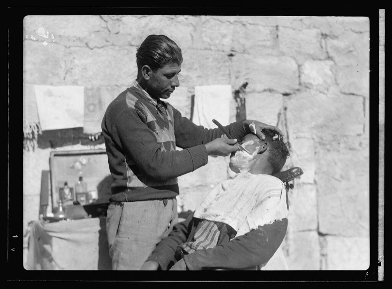 A street barber circa 1934-39 wears high-waist trousers with a v-neck sweater. 
