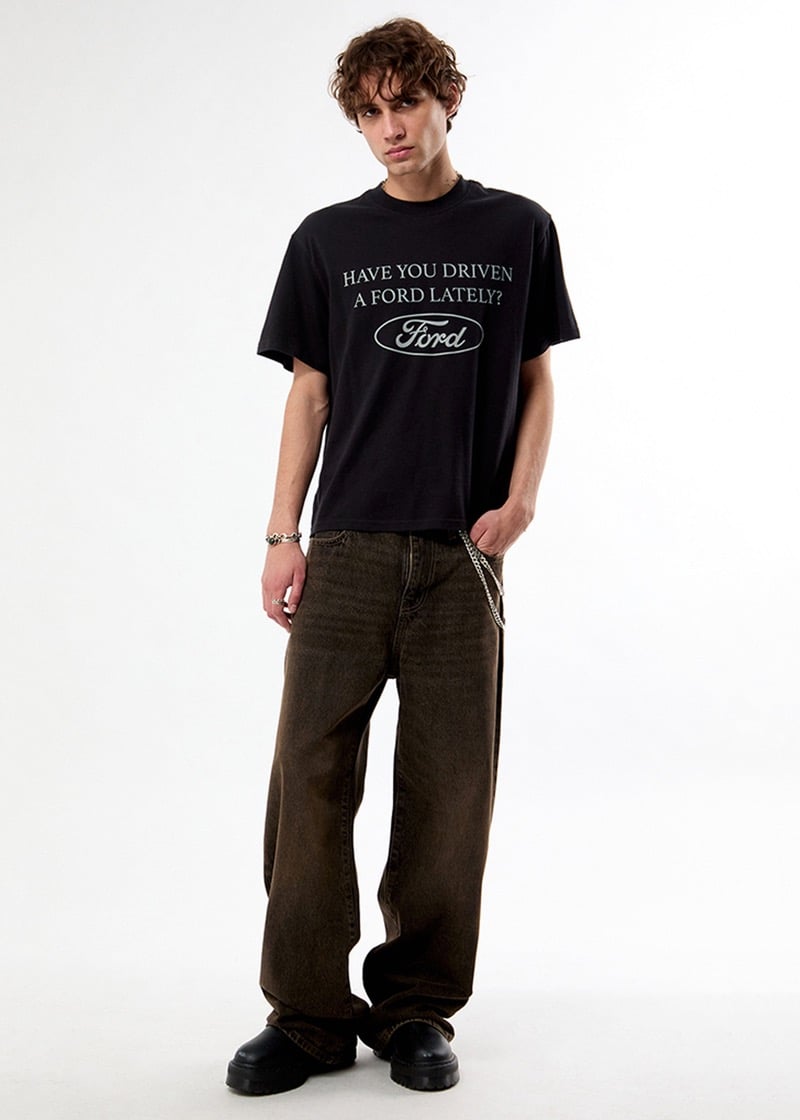 Man wearing a black “Have You Driven a Ford Lately?” graphic T-shirt with dark faded jeans, a wallet chain, and black shoes, standing against a plain background.