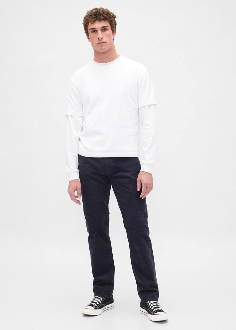 Man wearing a white short-sleeve T-shirt layered over a white long-sleeve shirt, paired with dark straight-leg jeans and black Converse sneakers, standing against a plain background.