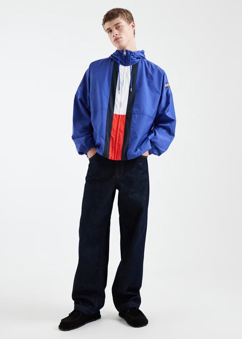 Man wearing a blue windbreaker with white and red paneling, paired with dark wide-leg jeans and black shoes, standing with hands in pockets against a plain background.
