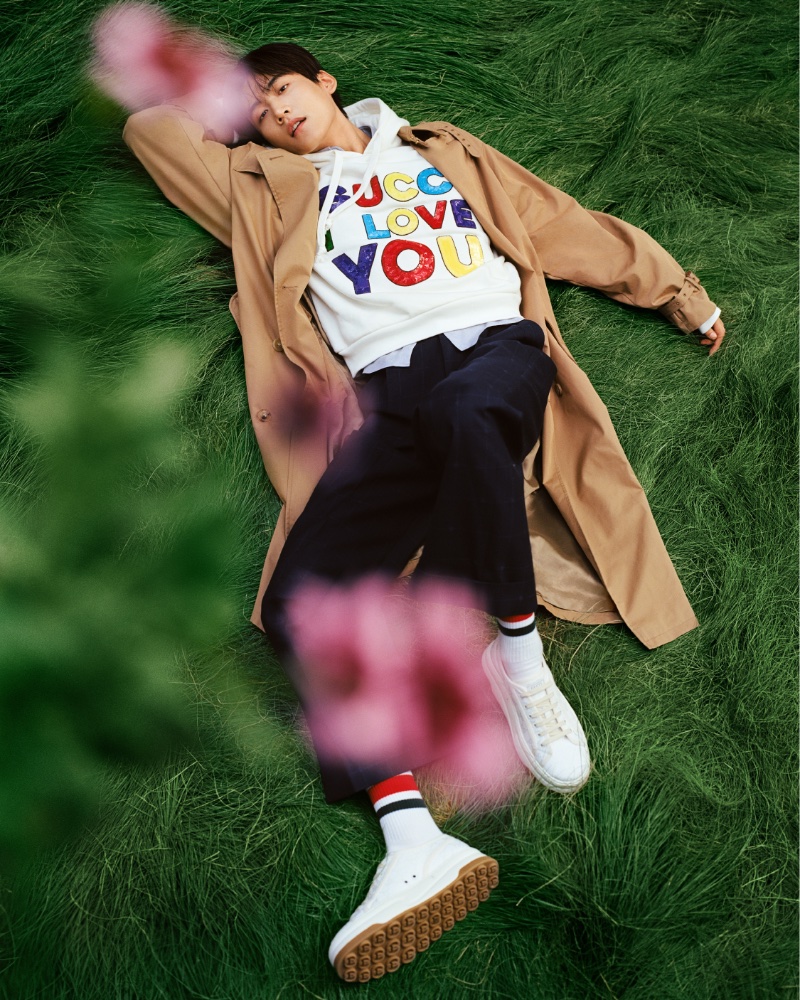 Laying in the grass, Daniel Zhou wears an oversized trench coat with a hoodie for Gucci's most recent men's campaign. 