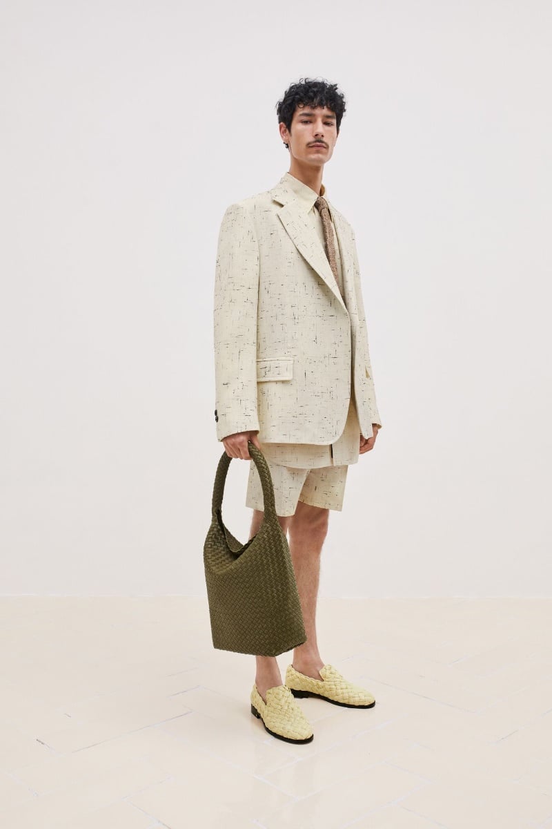 Noah Ramos cuts a dashing figure in a flecked cream short suit with a coordinated tie, accessorized with woven loafers and an olive tote, embodying Bottega Veneta's luxurious yet understated style. 