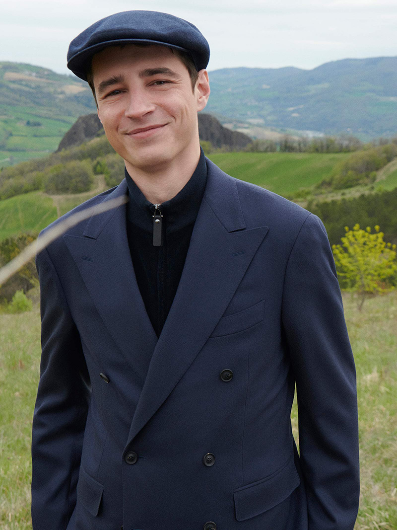 Adrien Sahores exudes charm in a navy double-breasted coat and a classic flat cap, set against the serene backdrop of rolling hills.