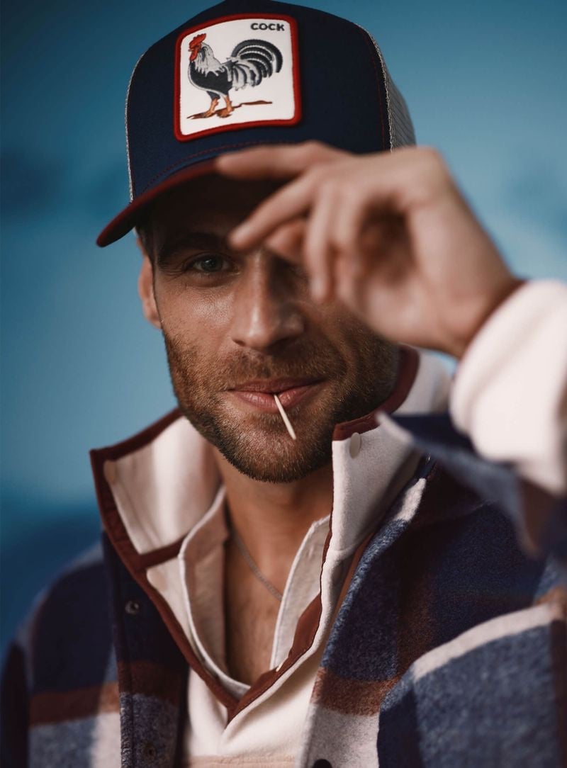 George Alsford strikes a casual pose, tipping his 'rooster' emblazoned cap with a playful glance, clad in a plaid-lined jacket from Simon's Le 31 collection.