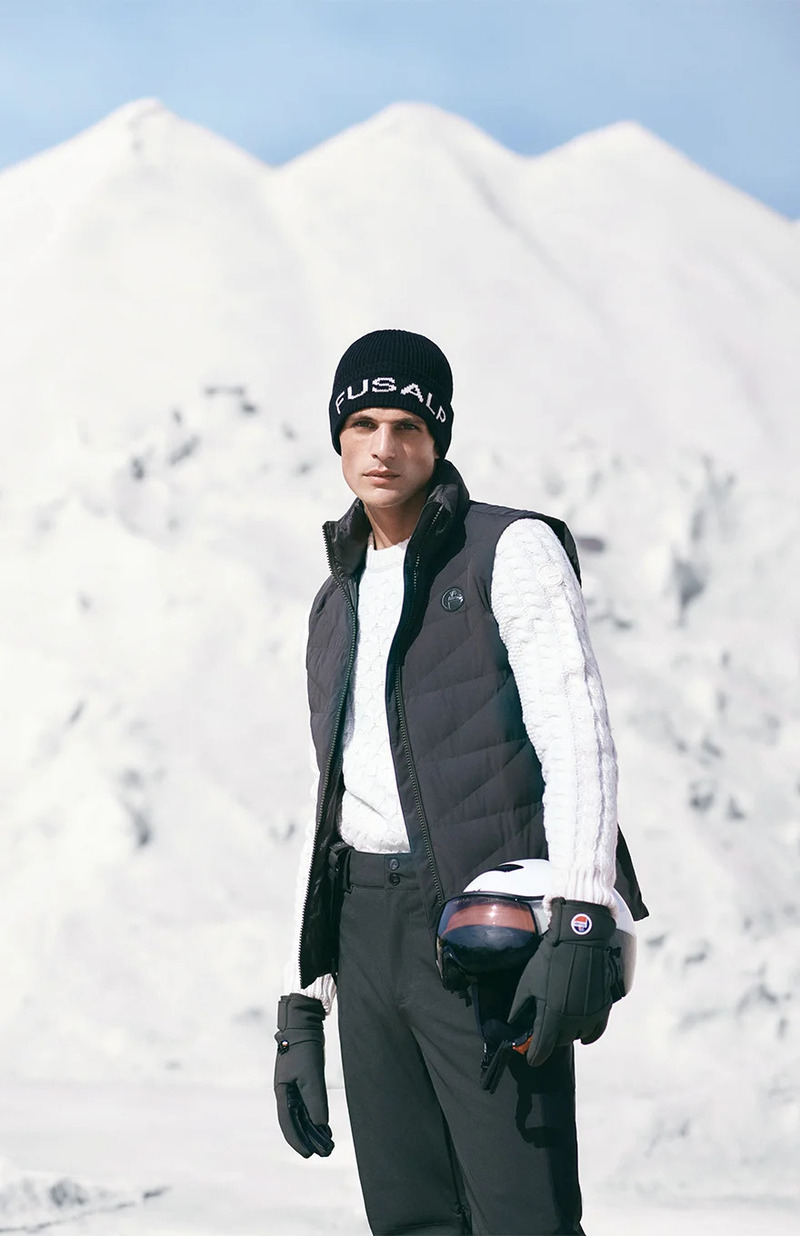 Pau Ramis stands against a snowy mountain backdrop, clad in a chic quilted vest and cable-knit sweater, complete with a Fusalp beanie and ski helmet in hand.