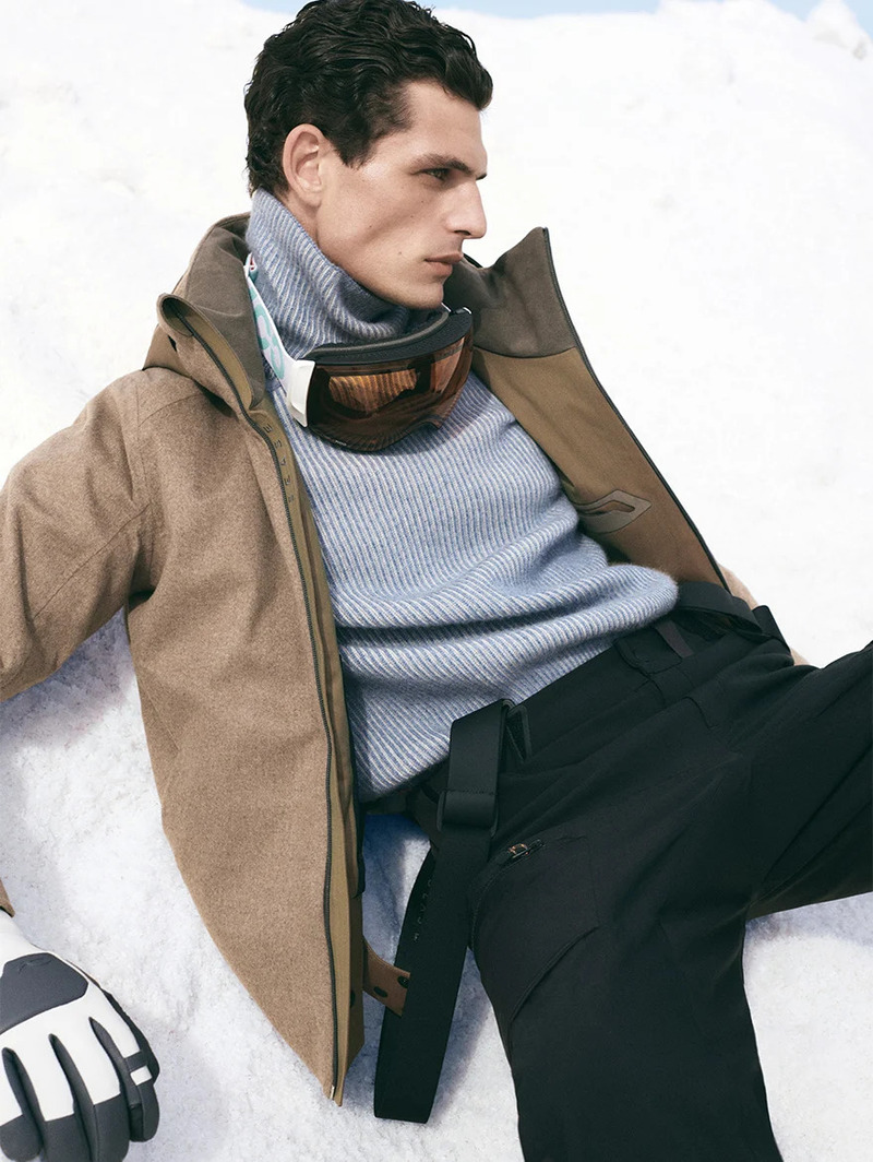 Pau Ramis lounges in the snow in a layered ensemble of a ribbed turtleneck and a taupe jacket, with Kask ski goggles resting on his collar.