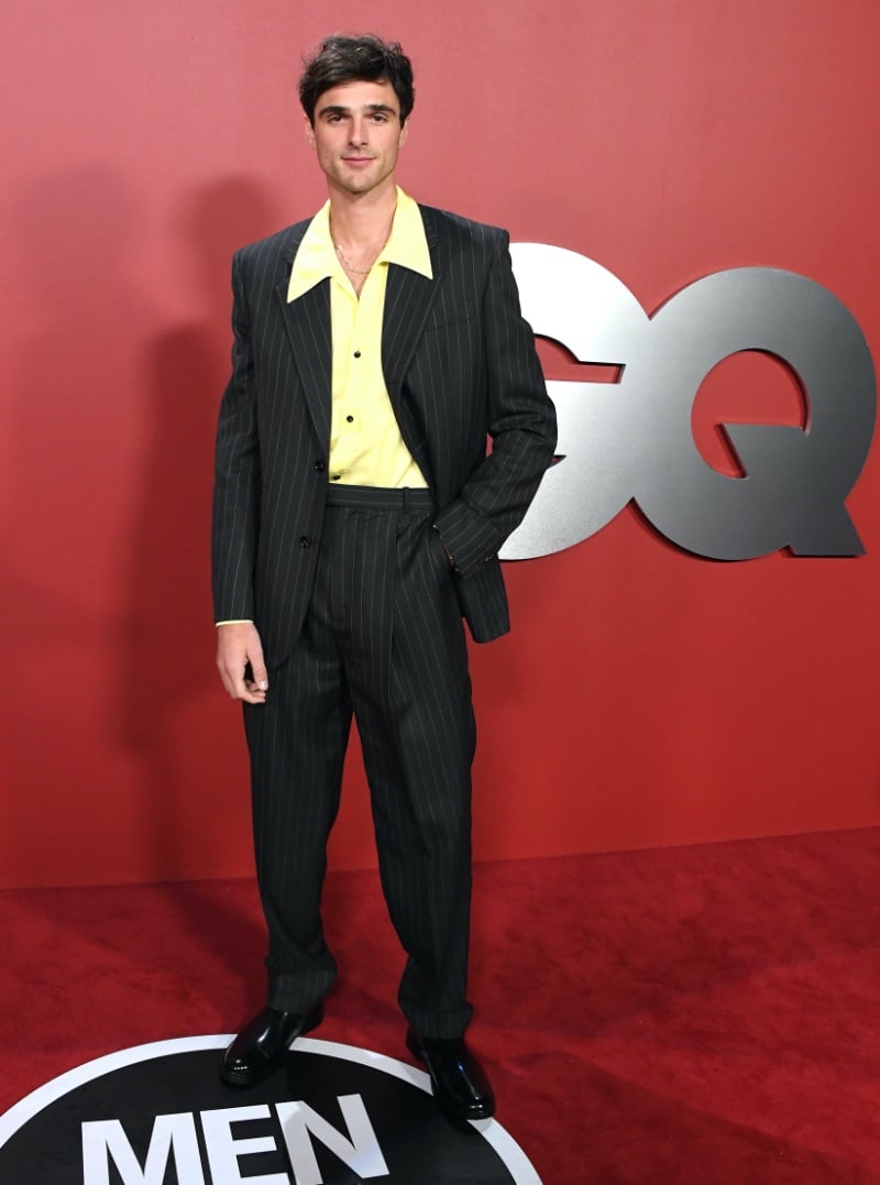 Jacob Elordi makes a bold statement at the 2023 GQ Men of the Year Awards, in a custom Bottega Veneta dark grey pinstripe wool suit, featuring a three-button single-breasted jacket with wide-leg trousers, and a vibrant pineapple-hued fluid viscose twill shirt that adds a pop of retro flair.
