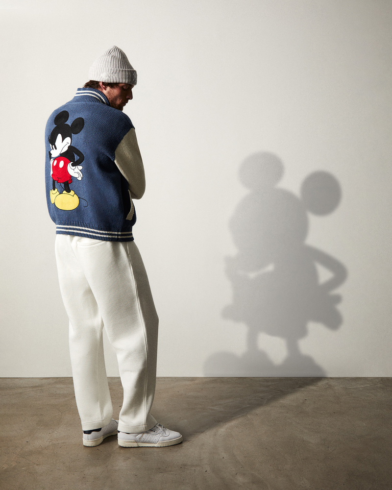 Rocky Harwood stands in profile, donning a cozy grey beanie with a Mickey Mouse knit jacket from the Kith for Mickey & Friends collection.