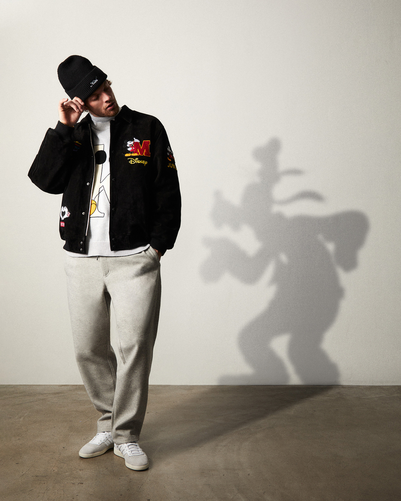 Model Rocky Harwood cuts a casual pose in a black varsity jacket featuring Disney motifs, his shadow casting a playful Goofy silhouette on the wall beside him.