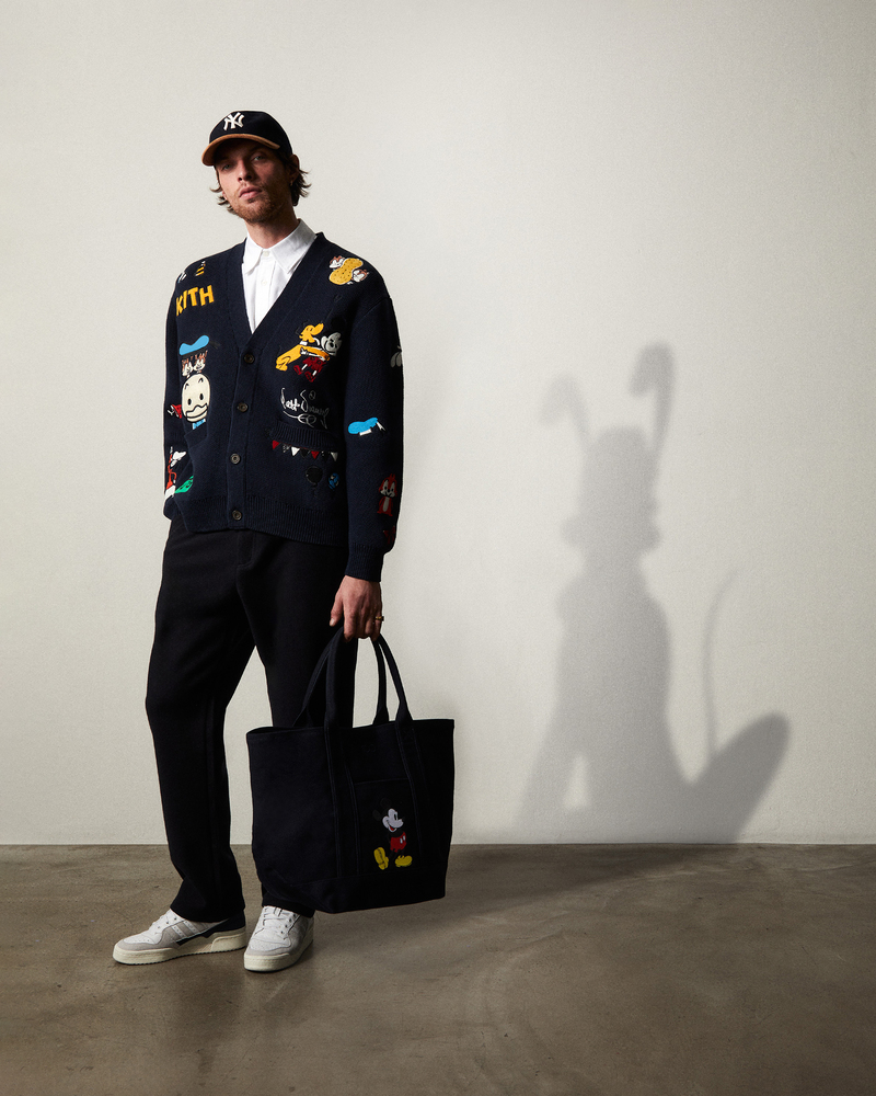 Rocky Harwood projects effortless style in an embroidered cardigan with Disney characters, complemented by a classic baseball cap and a thematic tote bag. 