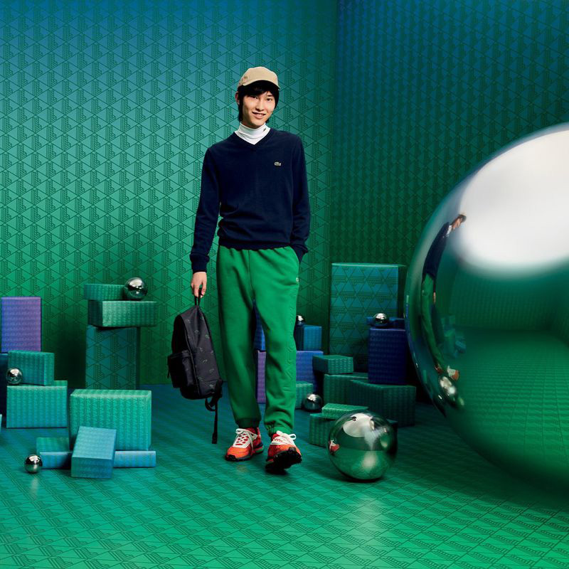 Hyojin Jung stands out in a vibrant ensemble, pairing a navy Lacoste V-neck sweater over a crisp white turtleneck, with striking green pants and a cap, all set against an abstract geometric backdrop for Lacoste's Holiday 2023 campaign.