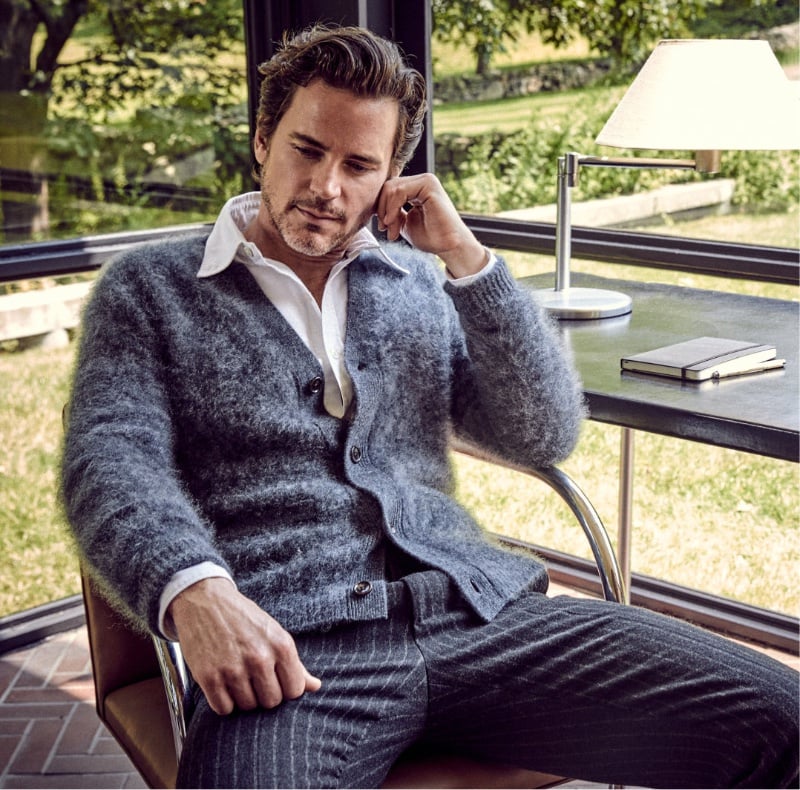 Matt Bomer, deep in thought, sits comfortably in a luxurious brushed cashmere cardigan and pinstripe suit pants, complementing a timeless poplin shirt.