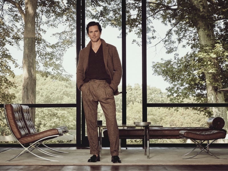Standing confidently amidst modern architecture, Matt Bomer is the epitome of sophistication in a Todd Snyder ribbed Donegal V-neck sweater, Madison sport coat, and suit pants, finished with elegant tassel loafers.